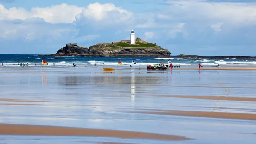 This landscape photograph shows Godrevy Beach in Cornwall, England, during a summer morning. The main subject of the image is Godrevy surfing at Gwithian, with several surfers and beachgoers visible along the shallow shoreline where the wet sand creates clear reflections of the people and flags. In the background, the sea meets Godrevy Island, which features the prominent Godrevy Lighthouse, a well-known landmark of the Cornwall coastline in the United Kingdom. The calm summer light and blue skies enhance the vivid colours typical of the region, while the gentle waves underline the beach's popularity as a surfing destination.